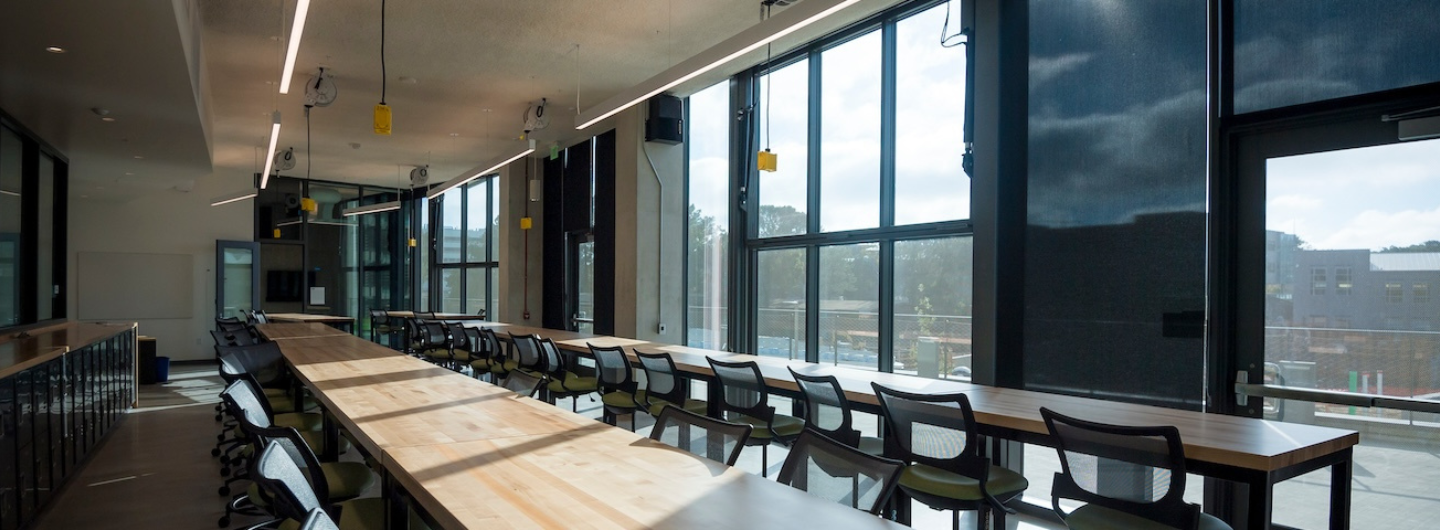 alt="A large, empty room with long wooden tables and black chairs, featuring floor-to-ceiling windows along one side, offering a view of the outside"